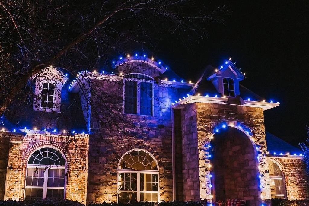 Two-story stone and brick home in Northwest Arkansas lit with blue and warm white C9 Christmas lights along every roofline and arch