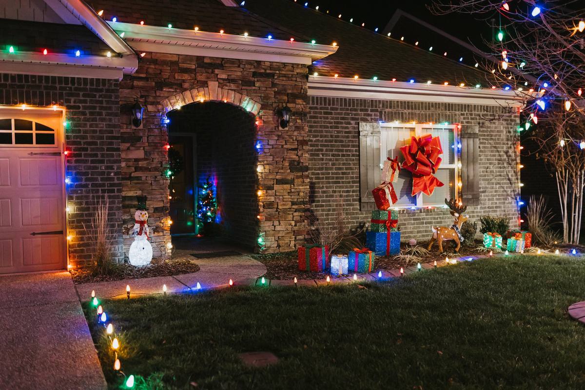 Brick home entry decorated with multicolor C9 Christmas lights, lit gift boxes, a snowman, a reindeer and a lit walkway