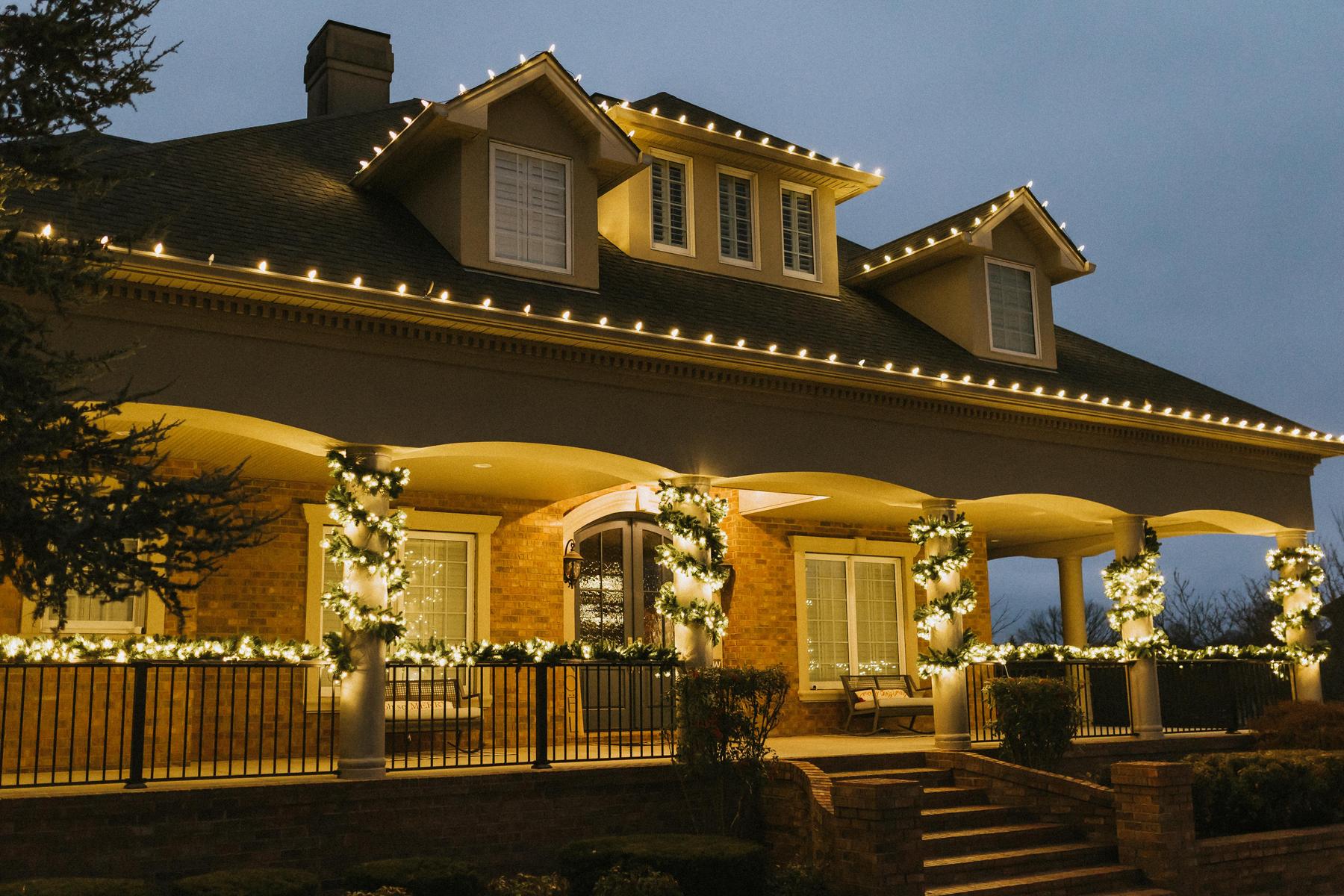 Large brick home in Bentonville, Arkansas outlined in warm white C9 Christmas lights with lit garland wrapped around the front porch columns at dusk