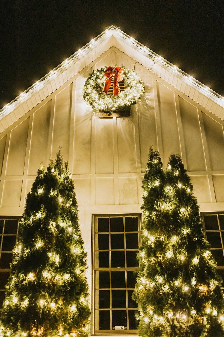 White gabled home with a large lit wreath and two illuminated evergreen trees flanking the entry